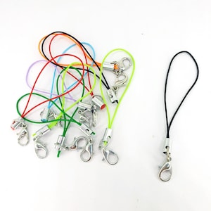 Può includere: Assortimento di ciondoli per telefono colorati con chiusure argentate. I ciondoli sono realizzati in cordoncino sottile e hanno una perla argentata all'estremità. Un ciondolo è nero.