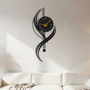 Puede incluir: Reloj de pared negro y dorado con diseño moderno. El reloj tiene una esfera circular con manecillas doradas y fondo negro, enmarcado en una estructura abstracta de metal negro. Montado en una pared blanca sobre un sofá blanco.