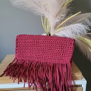 Può includere: Pochette all'uncinetto bordeaux con frange. La borsa rettangolare è realizzata con un filato spesso e strutturato e presenta lunghe frange lungo il bordo inferiore. La borsa è esposta su un tavolo bianco.