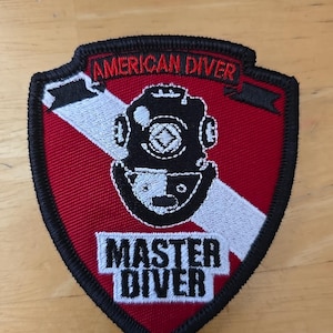 Puede incluir: Un parche bordado en forma de escudo con fondo rojo y una franja blanca diagonal. Presenta un gráfico de casco de buzo negro, el texto AMERICAN DIVER en la parte superior y el texto MASTER DIVER en la parte inferior, todo bordeado en hilo negro.