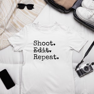 Könnte beinhalten: Weißes T-Shirt mit dem Text "Shoot. Edit. Repeat." in Schwarz bedruckt. Das Shirt liegt flach auf einer weißen Oberfläche mit einer Kamera, einer Sonnenbrille und einem Koffer im Hintergrund.