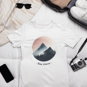 May include: White t-shirt with a watercolor design of mountains and a sunset in a circle. The text "Rise Above" is printed below the design.