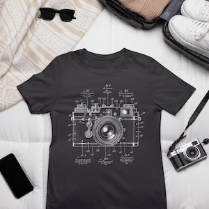 May include: A dark gray t-shirt with a white graphic of a camera patent drawing. The drawing includes numbers and text describing the camera's parts.
