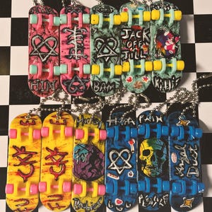 May include: A collection of miniature skateboard keychains in various colors. Each keychain features unique graffiti-style artwork, including hearts, skulls, and text. The skateboards have pink, yellow, blue, and green wheels and trucks, and are attached to silver chains.