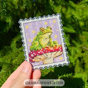 May include: A green frog sits on a red and yellow mushroom with a purple background. The image is a cross-stitch pattern.