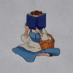 Op de afbeelding: Kruissteek van een persoon die een blauw boek leest, met een bruine mand. De persoon draagt een blauwe jurk en een witte blouse. De kruissteek staat op een witte achtergrond.