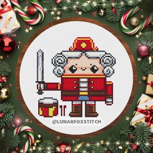 Op de afbeelding: Een kruissteekontwerp van een cartoon notenkraker soldaat in een rode jas en hoed, met een zwaard. Het ontwerp staat in een witte cirkel, omringd door kerstversieringen. De tekst "@LUNARFOXSTITCH" staat onderaan.