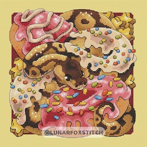 May include: Cross-stitch design featuring a snake coiled around colorful donuts. The donuts have pink, white, and brown frosting with sprinkles. The snake is brown and tan with a dark brown head. The background is a light yellow color. Text reads "@LUNARFOXSTITCH".