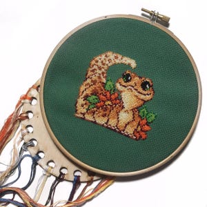 May include: A cross-stitch embroidery hoop with a green background featuring a brown and tan gecko with orange flowers. The hoop is surrounded by a variety of colourful embroidery floss.