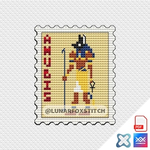 May include: A cross-stitch pattern shaped like a postage stamp featuring the Egyptian deity Anubis in black, blue, yellow, and brown. The text ANUBIS is printed vertically in red on the left, with @LUNARFOXSTITCH at the bottom on a tan background.