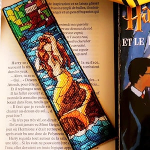 May include: A colorful bookmark featuring a stained glass style image of a mermaid with a yellow tail and blue and green water. The bookmark is placed on top of a book with the title "Harry Potter and the ..." visible.