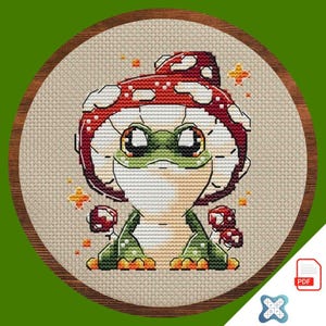 May include: Cross-stitch embroidery of a whimsical frog wearing a red and white mushroom cap. The frog is green with large eyes and is surrounded by small mushrooms and starburst accents. The embroidery is set in a round wooden frame.
