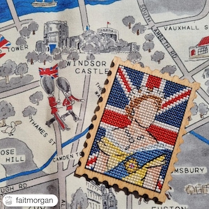Puede incluir: Broche de madera con forma de sello postal, con un diseño de punto de cruz de una mujer con corona sobre una bandera de la Union Jack. El broche descansa sobre una tela tipo mapa con ilustraciones del Castillo de Windsor.