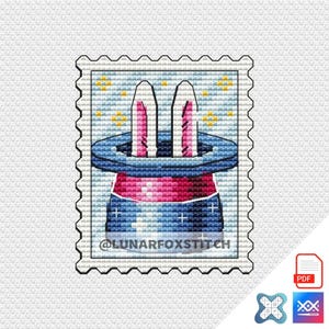 May include: A cross-stitch pattern shaped like a postage stamp featuring a dark blue magician's top hat with a bright pink band. Two white rabbit ears with pink inner details emerge from the hat against a light blue background with yellow stars.