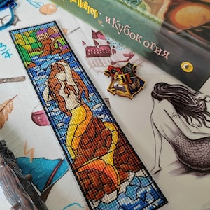 May include: A rectangular cross-stitch bookmark featuring a stained-glass style design of a mermaid with a yellow tail and brown hair. The artwork includes blue water and brown rock elements, finished with a black border and a detailed stitch pattern.