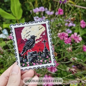 May include: A black crow perched on a tombstone in a graveyard, with a full moon and cross in the background. The image is a cross-stitch design on a red, white, and black fabric.