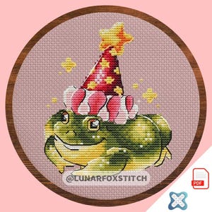 May include: Cross-stitch design of a green frog wearing a party hat with a gold star. The frog is smiling, and the design is set in a round wooden frame. The text "@LUNARFOXSTITCH" is at the bottom. The background is a light pink fabric.