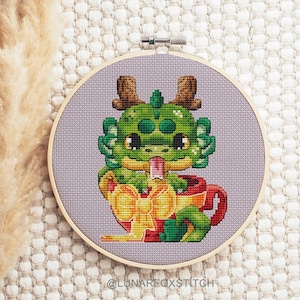 May include: A green dragon with a red and yellow bow is sitting in a red teacup. The dragon has a pink tongue sticking out and is looking to the right. The teacup has a white handle.