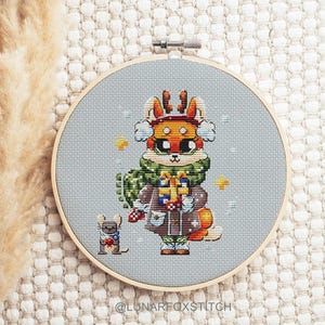 Puede incluir: Patrón de punto de cruz de un zorro con una bufanda verde y sosteniendo un regalo, con un pequeño ratón con una bufanda a su lado. El fondo es de color azul claro con copos de nieve.