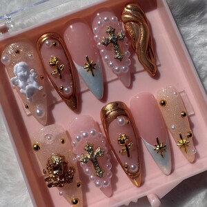May include: A set of ten pink and white press-on nails with gold accents. The nails feature pearl beads, gold crosses, gold roses, and gold angel wings.