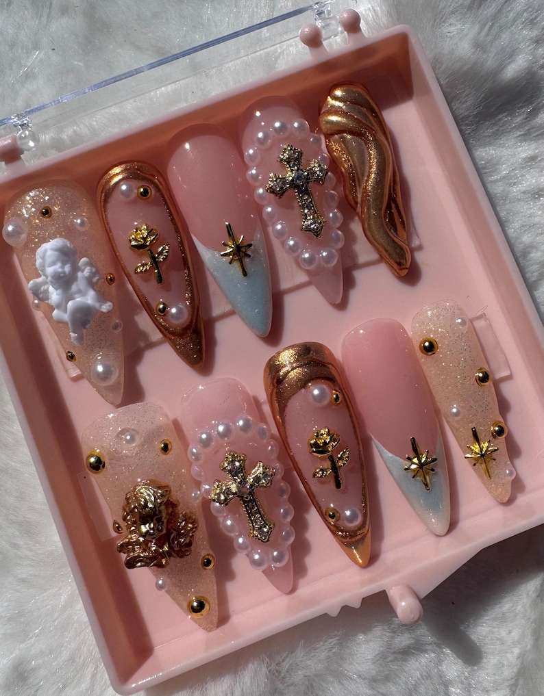 May include: A set of ten pink and white press-on nails with gold accents. The nails feature pearl beads, gold crosses, gold roses, and gold angel wings.