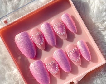 May include: A set of ten pink glitter press-on nails with a pearl heart design. The nails are in a clear plastic box with a pink lid.