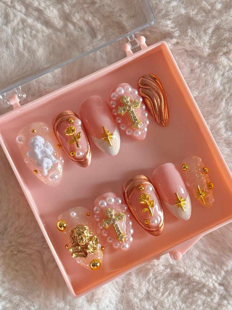 May include: A set of pink press-on nails with gold accents. The nails feature a variety of designs, including a cross, a rose, an angel, and a wing. The nails are arranged in a pink box.