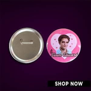 May include: Pink button with a picture of a man with brown hair and brown eyes. The button has a heart shape with a butterfly and the text "Team Edward!"