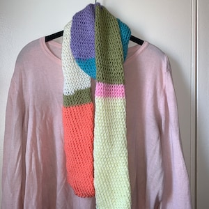 May include: A colorful knitted scarf with stripes of purple, teal, green, pink, and orange. The scarf is hanging on a hanger against a white wall.