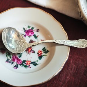 May include: A silver ladle with an ornate design sits on a white plate with a floral pattern. The plate has a gold rim.