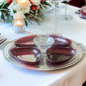 May include: A silver serving tray with four burgundy-colored dishes and a small clear glass bowl in the center. The tray has a ribbed design and handles. The dishes are kidney-shaped. The setting is on a white tablecloth.