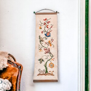 May include: A vintage embroidered wall hanging with a floral design featuring a variety of colorful flowers and leaves on a white background.