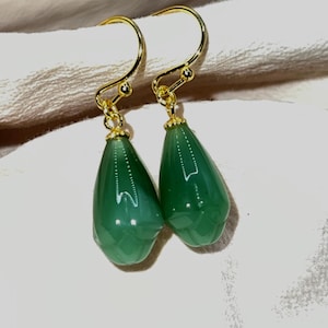 Peut inclure: Une paire de boucles d'oreilles à crochet dorées avec des pendentifs en forme de goutte d'eau en pierres précieuses vertes translucides. Les boucles d'oreilles sont présentées sur un fond en tissu de ton neutre.