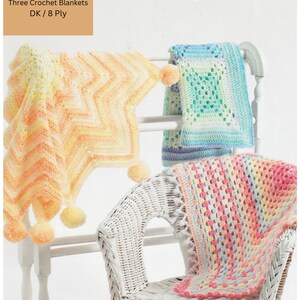 May include: Three crochet baby blankets in pastel colors, with pom-poms and fringe. The blankets are displayed on a white wicker chair and a white wooden rack. The text "Three Crochet Blankets DK/8 Ply" is visible at the top of the image.