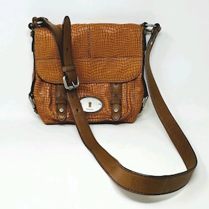 以下が含まれることがあります： 茶色の革製クロスボディバッグ。クロコダイルのような質感の模様があります。フラップ開閉、調節可能な長いストラップ、シルバーカラーの金具付き。前面にFossilのロゴが見えます。普段使いにデザインされています。