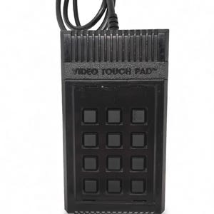 May include: A black rectangular Video Touch Pad controller featuring a grid of twelve square buttons. The device has a ribbed top section and a black power cord extending from the top, secured with a small grey cable tie for organization.