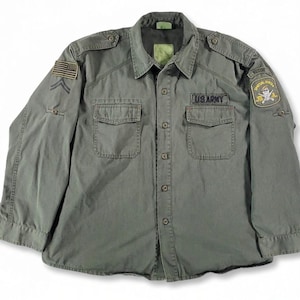 Könnte beinhalten: Olivgrünes Hemd im Militärstil mit Knopfleiste, zwei Brusttaschen und Schulterklappen. Mit einem US-Flaggen-Aufnäher, einem "US ARMY"-Aufnäher und einem "SPECIAL FORCES"-Aufnäher. Das Hemd hat lange Ärmel und einen Kragen.