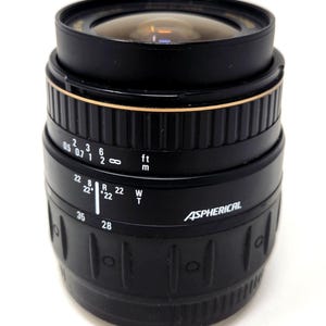 May include: A black camera lens with gold detailing and a clear glass lens. The lens features measurement markings in feet and metres, with the word "Aspherical" printed on the side. Designed for photography.