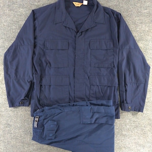 Puede incluir: Una camisa utilitaria de manga larga en color azul oscuro con cuello en punta, cierre frontal de botones y cuatro grandes bolsillos rectangulares de parche en la parte delantera. Debajo hay un pantalón azul oscuro a juego con un clip metálico.