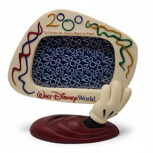 May include: A cream-colored Walt Disney World picture frame featuring the text 2000 and Celebrate the Future Hand in Hand. The frame is supported by a cartoon hand emerging from a dark red base, with colorful streamers decorating the border.