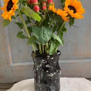 Könnte beinhalten: Eine schwarze Keramikvase mit einer texturierten, weiß gesprenkelten Oberfläche, gefüllt mit einem Strauß aus leuchtend gelben Sonnenblumen, grünen Blättern und kleinen runden roten Beeren. Die Vase hat eine unregelmäßige, organische Form.