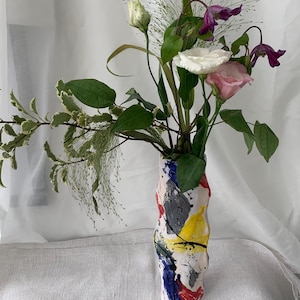 Puede incluir: Un jarrón de cerámica colorido con un diseño abstracto, que contiene un ramo de flores. El jarrón presenta salpicaduras de pintura roja, amarilla, azul y gris. Las flores incluyen flores blancas, rosas y moradas, con follaje verde.