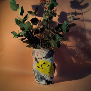 Könnte beinhalten: Eine Keramikvase mit einem abstrakten Design in den Farben Rosa, Schwarz, Weiß und Gelb. Die Vase ist mit einem Strauß grüner Eukalyptusblätter und roten Beeren gefüllt.