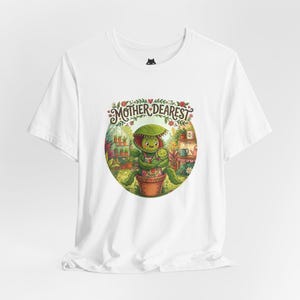May include: White t-shirt with a vibrant illustration of a plant-like character, labelled "Mother Dearest." The design depicts a plant-like figure embracing a smaller plant in a pot, set against a backdrop of a greenhouse and flowers.