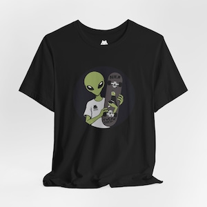 May include: A black short-sleeve t-shirt featuring a graphic of a green alien wearing a white shirt and holding a grey skateboard. The skateboard has the text ALIEN and TWERK SHOP printed on it. A small white cat logo is visible on the neck label.