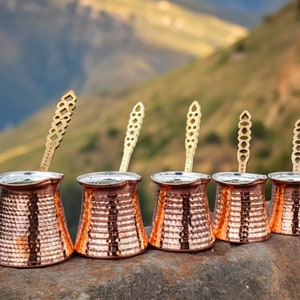 May include: A collection of five copper coffee pots, each with a hammered texture and a brass handle. The pots are arranged in a row, with the handles extending upwards. The background features a blurred view of a mountain landscape.