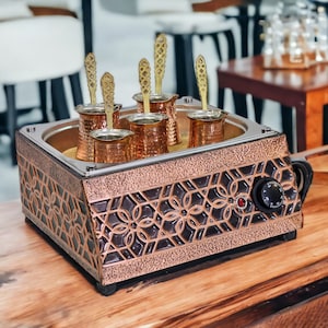 以下が含まれることがあります： 6つの個別のポットと温度調節ノブが付いた銅と真鍮製の電気コーヒーメーカー。コーヒーメーカーは木製の表面にあります。