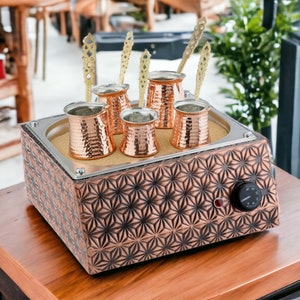 Op de afbeelding: Een elektrisch verwarmingsbakje van koper met een decoratief stervormig patroon. Vijf kleine koperen koffiekannen met handvatten staan op het bakje. Het bakje heeft een knop om de temperatuur in te stellen.