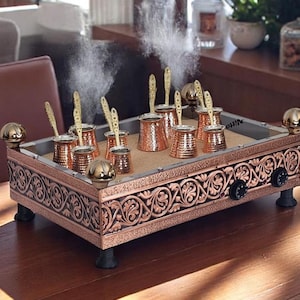Puede incluir: Una cafetera de color cobre con varias ollas pequeñas y cucharas, colocada sobre una base rectangular decorativa. La base tiene tallas ornamentadas y está soportada por patas cortas. El vapor se eleva de las ollas, lo que sugiere que están en uso.