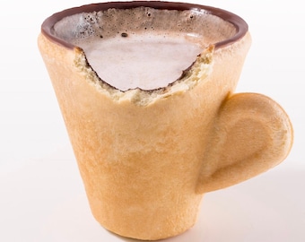 May include: A brown edible cup with a chocolate rim filled with a light brown liquid. The cup has a bite taken out of the rim and a handle.